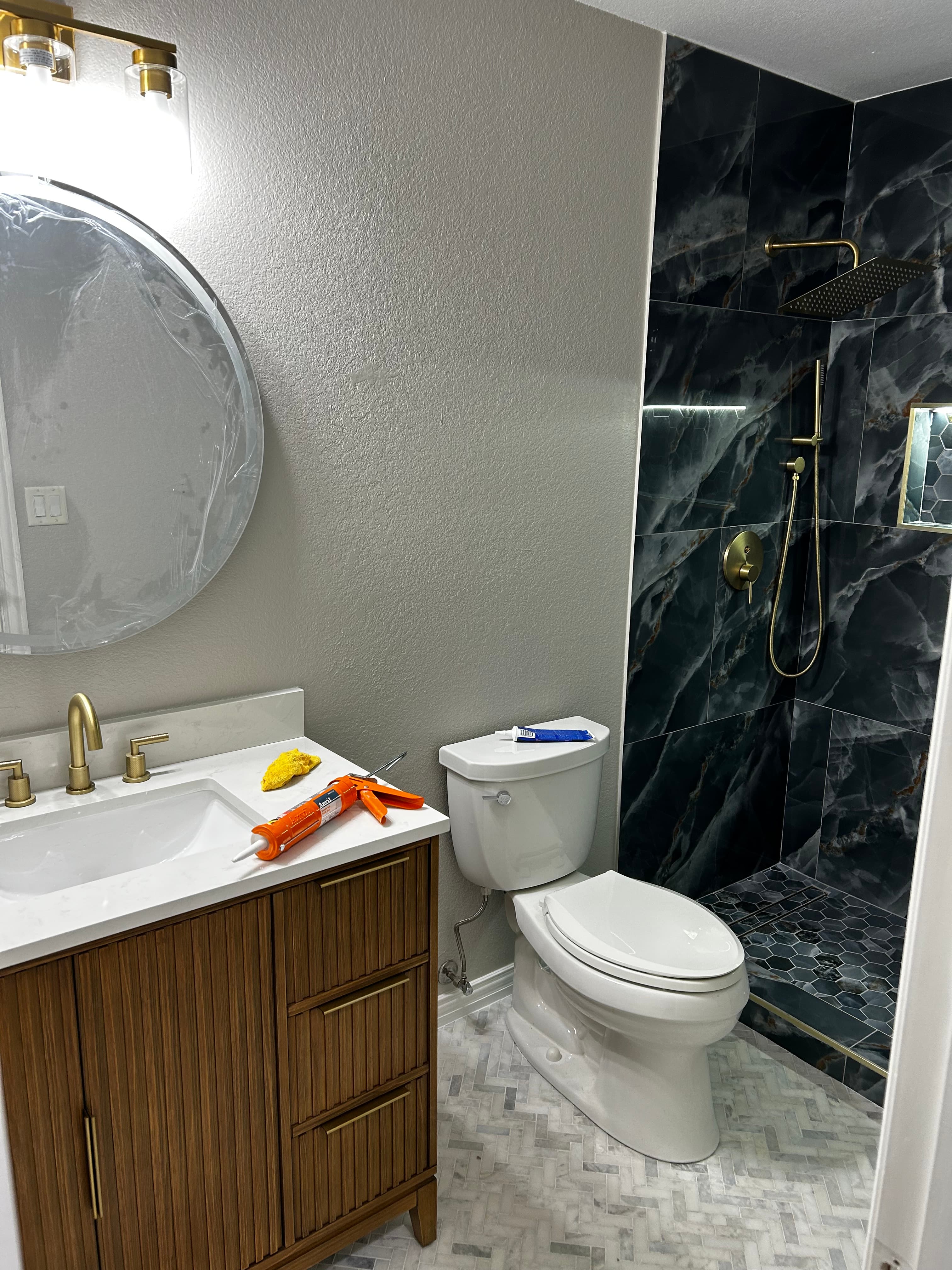 Bathroom remodel with dark marble-look shower tile, brushed-gold fixtures, a wood-slat vanity, and a round mirror