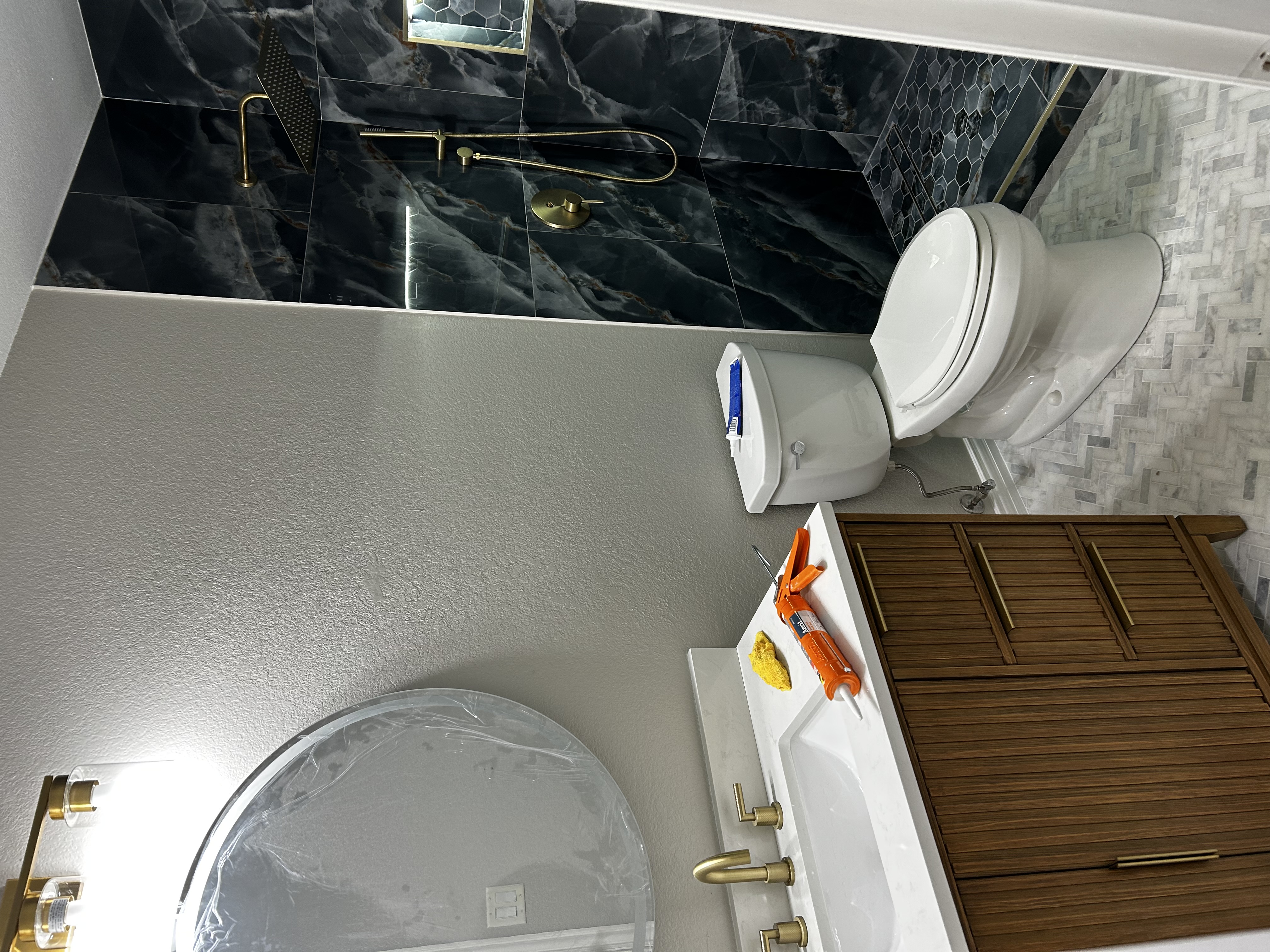 Bathroom remodel with dark marble-look shower tile, brushed-gold fixtures, a wood-slat vanity, and a round mirror